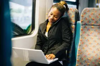 Eine Frau sitzt telefonierend mit ihrem Laptop im Bus und arbeitet.