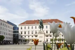 Das Reiterdenkmal auf dem Wittelsbacherplatz im Sommer. Im Vordergrund sind blühende Blumen zu sehen.