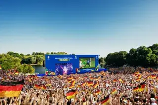 Visualisierung der Fan Zone im Olympiapark: Deutsches Publikum mit Fahnen vor Großbild-Leinwand bei Sonnenschein