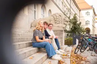 Schülerin und zwei Schüler sitzen auf den Stufen vor dem Maximiliansgymnasium in München