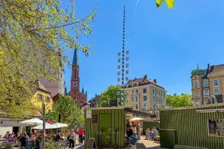 Interimsmarkt am Wiener Platz in München-Haidhausen mit grünen Marktmodulen, Podest, Maibaum und Kirchturm St. Johann Baptist im Hintergrund, April 2026