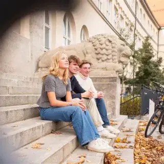 Drei Schülerinnen und Schüler sitzen vor dem Maximiliansgymnasium in München