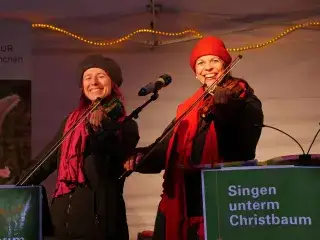 Monika Drasch (links) und Traudi Siferlinger (rechts) singen mit Geigen unterm Christbaum auf dem Münchner Christkindlmarkt