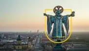 Blick auf das Münchner Kindl mit einem gelben Wappenrahmen über der Skyline Münchens